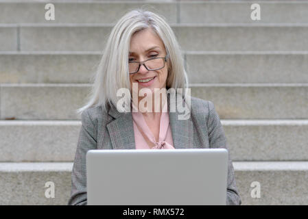 Attraente professional Donna che utilizza un computer portatile in città seduto su outdoor passi concreti di sorridere quando ella tipi Foto Stock