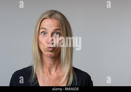 Stupito attraente donna bionda staring wide eyed alla telecamera mentre esercitano le sue labbra nella speculazione isolato sul grigio con spazio di copia Foto Stock