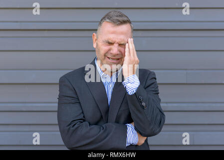 Sottolineato imprenditore, o che soffrono di mal di testa e piedi con la mano al suo tempio smorfie contro il grigio di una parete esterna Foto Stock