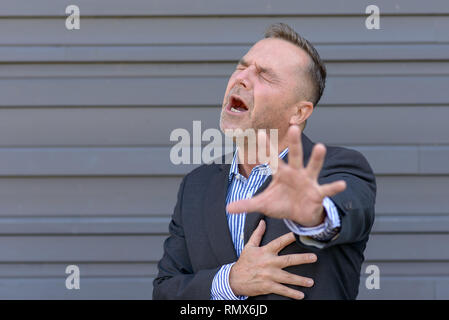 Imprenditore che impugnano il petto nel dolore o angoscia mentre raggiungendo fuori con il suo altro lato nella disperazione contro un muro grigio con spazio di copia Foto Stock