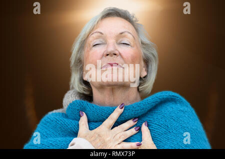 Attraente donna senior assaporando il momento in piedi con gli occhi chiusi e la testa inclinata indietro con un espressione serena come essa abbraccia il suo torace con h Foto Stock