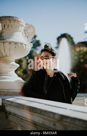 giovane donna di bellezza di moda posing in un parco che indossa occhiali una fontana sul retro Foto Stock