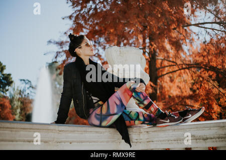 giovane donna di bellezza seduta in un recinto parco indossando occhiali guardare camera fontana indietro autunno giorno Foto Stock