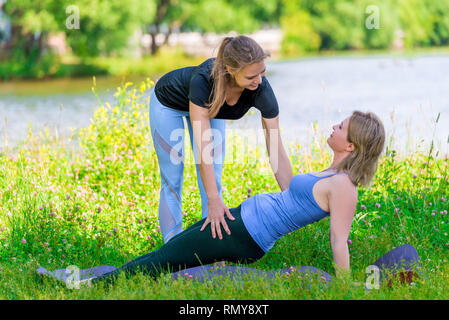 Trainer aiuta a eseguire correttamente gli esercizi di yoga per una donna matura, sport nel parco in estate Foto Stock