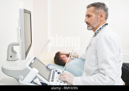 Grave medico seduto con il client nel gabinetto medico. Terapista facendo diagnostica ad ultrasuoni, guardando il monitor, tenendo la sonda. Specialista di esaminare i linfonodi sul collo. Foto Stock