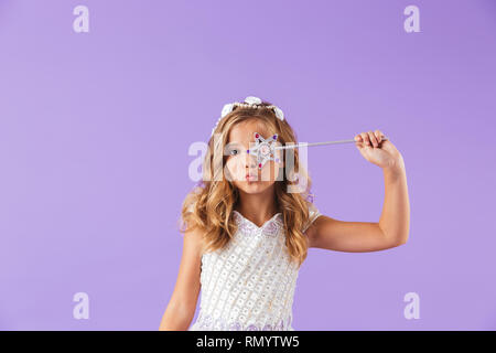 Ritratto di un sorridente carino Pretty girl vestito con un abito principessa isolate su sfondo viola, tenendo la bacchetta magica Foto Stock