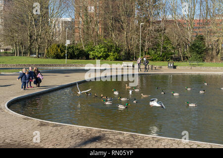 La famiglia (madre con tre bambini) dar da mangiare alle anatre e gabbiani nel Parco Eastrop, Basingstoke, UK, su una soleggiata giornata di febbraio Foto Stock