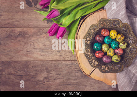 Foto scure di Pasqua uova di quaglia in un antico recipiente metallico. Bellissimi fiori di primavera - viola i tulipani su uno sfondo di legno. Cornice floreale con splendide Foto Stock