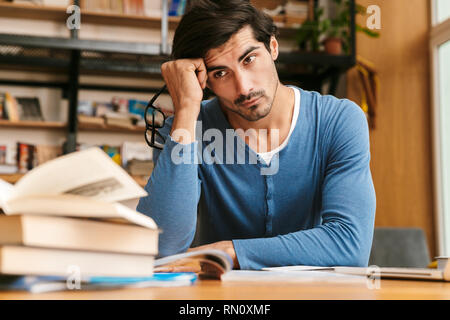 Bello stanco giovane uomo seduto al banco della libreria, lavorando/studio Foto Stock