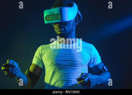 Di carnagione scura tourisistic maschio ventola con 3D tour in Asia, in piedi con head mounted display e controller palmare su sfondo scuro con rosso blu Foto Stock