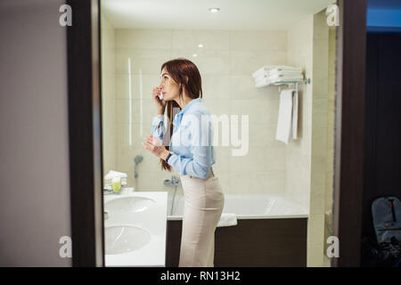 Immagine della bella femmina vestito in romantica blusa blu, gonna a matita guardando lo specchio mentre si lavano le mani al mattino su bianco lavello in b Foto Stock