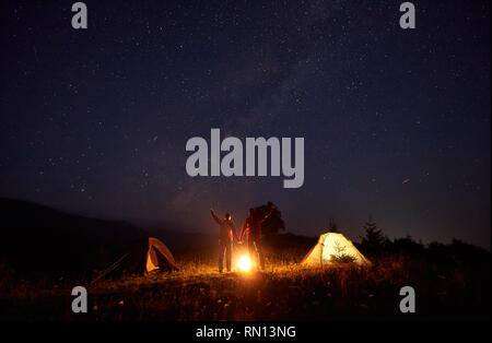 Bella notte campeggio foto. Luminose falò bruciando tra un ragazzo e una ragazza in piedi con le braccia alzate di fronte illuminata tende sotto scuro incredibile cielo stellato su lontane colline sullo sfondo. Foto Stock