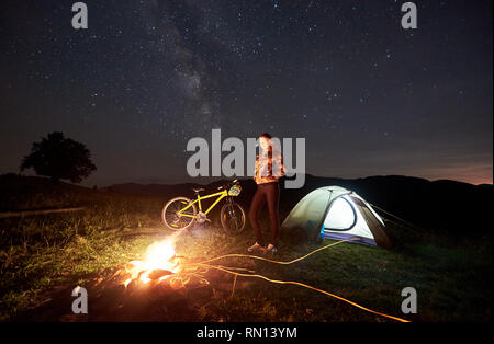 Giovane donna ciclista in appoggio di notte campeggio vicino a bruciare falò, illuminato tenda turistica, mountain bike serata sotto il cielo pieno di stelle e la Via Lattea. Attività all'aperto e il concetto di turismo Foto Stock