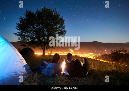 Vista posteriore di felice coppia romantica viaggiatori aventi un riposo a falò vicino illuminato blu tenda turistica sotto il cielo notturno pieno di stelle. Sullo sfondo il cielo stellato, montagne, grande albero e città luminosa Foto Stock