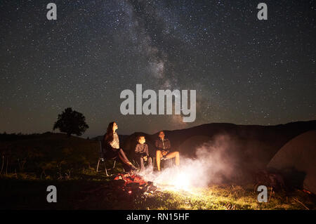 Giovane turista famiglia avente un insieme di riposo in montagna, seduti su sedie accanto a camp, falò e tenda a notte d'estate. Madre, padre e figlio godendo di notte il cielo stellato pieno di stelle e la Via Lattea Foto Stock