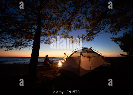 Il campeggio sulla riva del lago di notte. Tourist tenda sotto gli alberi e giovane famiglia, l uomo e la donna la preparazione di cibo sul fuoco sul blu scuro del cielo della sera e acqua chiara dello sfondo. Turismo e adventure concept. Foto Stock
