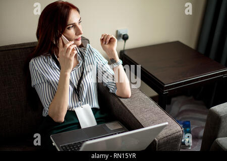Interessanti i capelli rossi femmina di project manager di marketing corporation indossando badge vuoto rendendo il lavoro a distanza seduta alla camera in hotel utilizzando laptop e Foto Stock