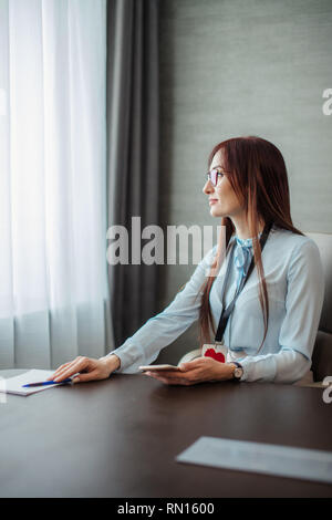 Malinconici donna commercialista con lunghi capelli allo zenzero, indossa occhiali si siede al suo posto di lavoro alla fine della giornata di lavoro e guardando la finestra dreamily, Foto Stock