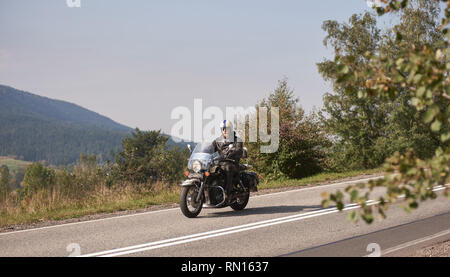 Barbuto biker in casco, occhiali da sole e pelle nera di equitazione abbigliamento cruiser potente moto verso il basso sunny strada asfaltata sulla luminosa giornata estiva sullo sfondo della verde collina di woody. Foto Stock