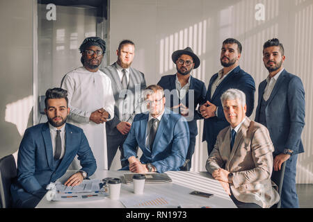 Bello caucasian mid-imprenditore invecchiato vestito come arabo si siede nella poltrona circondato con gruppo di formalmente vestiti business people, posa sul backg bianco Foto Stock