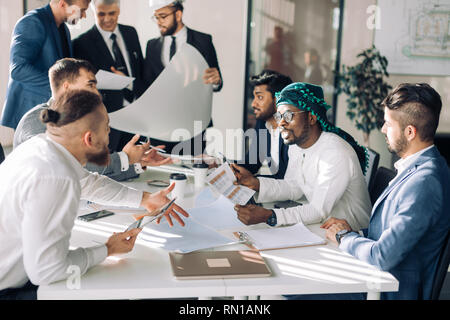 Gruppo multirazziale di costruttori, costruttori, genieri e architetti discutendo blueprint presso l'ufficio. Tutti gli uomini sono vestiti in abiti aziendali, e bianco Foto Stock