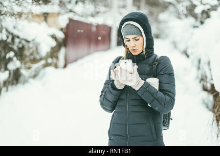 Bella Donna vestita di un nero giacca invernale con il telefono cellulare e termos sotto il suo braccio. Snow Landscape sfondo dietro Foto Stock