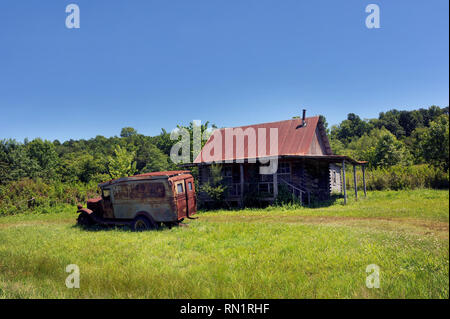 Vecchio jalopy siede nel cantiere di abbandono dei log cabin in Ozark Montagne dell'Arkansas. Cabina è dotata di tetto dello stagno e vecchio carrello è sbiadito cartello hillbilly. Foto Stock