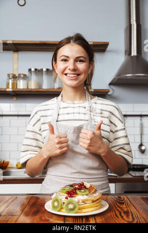 Immagine di una giovane e bella donna in cucina cucinare e mangiare la torta che mostra Thumbs up. Foto Stock