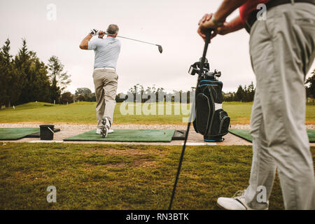 Senior man a una gamma di guida per giocare a golf e praticare il suo swing con un altro uomo in piedi di fronte. Senior golfisti maschio a fare pratica al campo da golf. Foto Stock