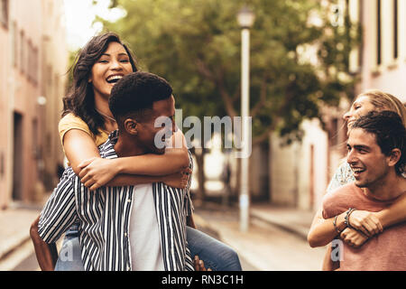 Due giovani uomini che trasportano un femmine amici loro indietro e sorridente. Gruppo di amici piggybacking all'aperto sulla strada della citta'. Foto Stock