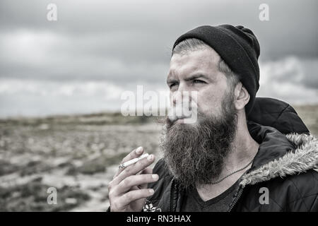 Barbuto hipster con barba lunga il fumo di sigarette in hat su sfondo naturale, cattiva abitudine e la tossicodipendenza, lo stile di vita Foto Stock