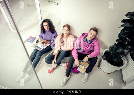 Tre studenti di indossare un abbigliamento informale seduti sul pavimento vicino classrom Foto Stock