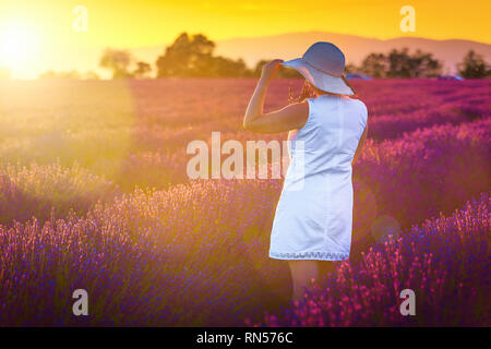 Allegro ragazza in un abito bianco e cappello di paglia per godersi la natura, campi di lavanda al tramonto. Paesaggio estivo con colorati viola i campi di lavanda Foto Stock