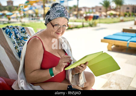 Senior donna in costume da bagno rosso in appoggio sulla sedia a sdraio su resort Foto Stock