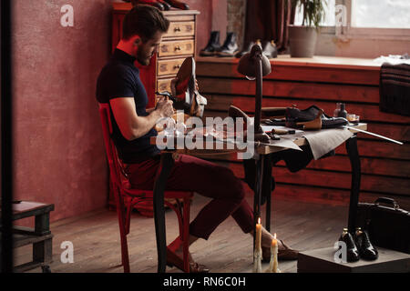 Giovane uomo slim tenendo un martello e un boot mentre è seduto al tavolo a lunghezza piena vista laterale foto Foto Stock