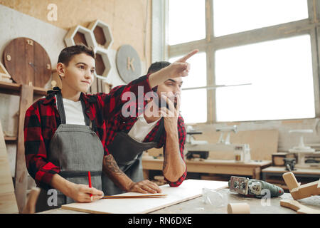 Famiglia, falegnameria, lavori in legno e concetto di persone - Maschio kid puntando con il dito a qualcosa e artigiano lo guarda in officina Foto Stock