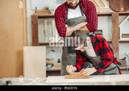 Carpenter guardando il suo piccolo partecipante lavora con giuntatrice al tavolo in legno con diversi strumenti di lavoro recante su di esso Foto Stock