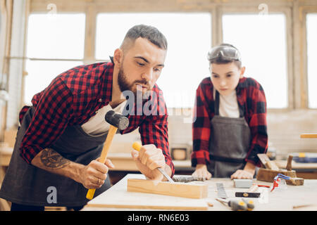 Giuntatrice barbuto uomo in occhiali di sicurezza e un grembiule lavora con uno scalpello in un negozio di falegnameria. Carpenter tenendo un bulino e suo figlio guardando il processo. Foto Stock