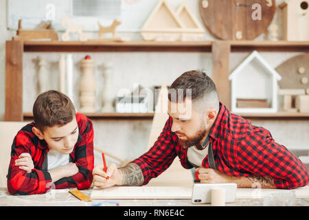 Papà che mostra il video su laptop a suo figlio prima di lavorare con il legno insieme, in piedi il falegname s workshop circondato con strumenti e figura in legno Foto Stock