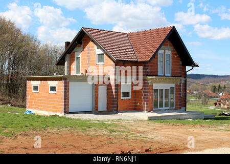Incompiuta in costruzione piccola suburbana di mattoni rossi casa di famiglia con annesso garage circondato con erba ed immondizia davanti con alberi di alto fusto Foto Stock