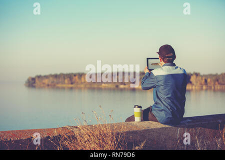 Un uomo si siede sul calcestruzzo argine di un lago pittoresco in serata. L'uomo prende una foto del lago con una compressa Foto Stock