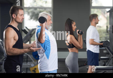 Vista laterale di persone che lavorano in una palestra moderna. Le donne e gli uomini utilizzando speciali attrezzature sportive. Trainer professionale aiutando, supporto di vecchio uomo facendo esercizi. Uomo anziano il sollevamento pesi. Foto Stock