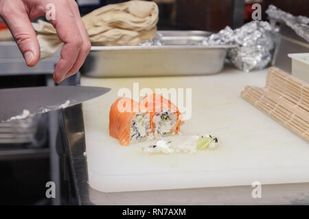 Primo piano della mano è il cuoco che taglia un rotolo di Philadelphia in un ristorante di cucina con un coltello affilato di sushi. Concetto di cucina piatti Giapponesi Foto Stock