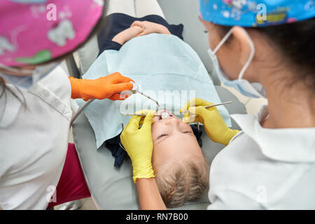 Dentisti lavorando con poco paziente in odontoiatria. KId giacente sulla poltrona del dentista con la bocca aperta. Stomatologists indossando i guanti, medico i cappucci e maschere. Gli specialisti che lavorano nella Clinica con bambino. Foto Stock