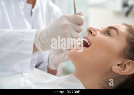 Primo piano della donna sdraiata sulla poltrona odontoiatrica con la bocca aperta e dentista in bianco guanti protettivi di esaminare i denti. Paziente di sesso femminile il trattamento e la cura sui denti. Concetto stomatologia e salute. Foto Stock