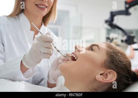 Stomatologia medico curante della donna di denti in odontoiatria clinica. Felice donna sdraiata con la bocca aperta. Dentista femmina in bianco uniforme e guanti utilizzando strumenti di ripristino. Foto Stock