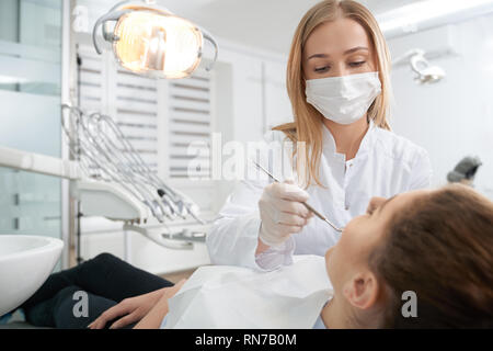 Professional dentista indossando maschera medica, guanti e bianco uniforme. Medico curante denti del paziente, impedendo la carie. Donna sdraiata in poltrona del dentista. Stomatologia concetto. Foto Stock