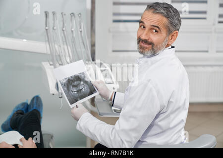 Professional, allegro medico di odontoiatria clinica seduto in poltrona. Barbuto, bello dentis guardando indietro alla fotocamera, sorridente e posa. Azienda Stomatologist apparecchiature odontoiatriche a raggi x in mani. Foto Stock