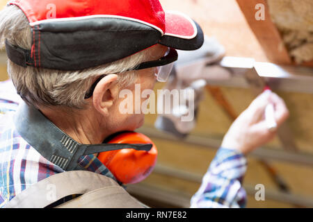 Uomo in googles facendo un segno sul telaio di metallo su attico incompiuto soffitto. Attico isolamento e rinnovo Foto Stock