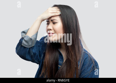 Ritratto di preoccupato bella bruna asian giovane donna casual in denim blu con camicia con il trucco permanente e disperata spiacenti tiene il capo coperto Foto Stock